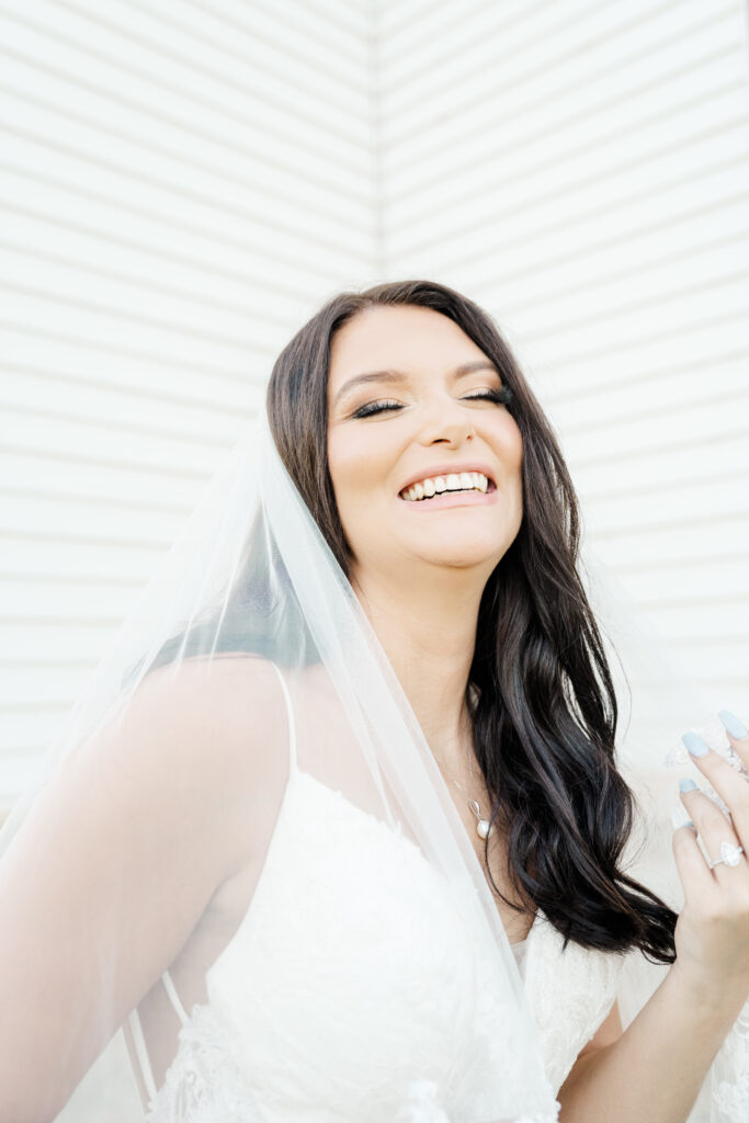 photo of a bride laughing in front of lubbock wedding venue, wearing a lace veil with light blue nails